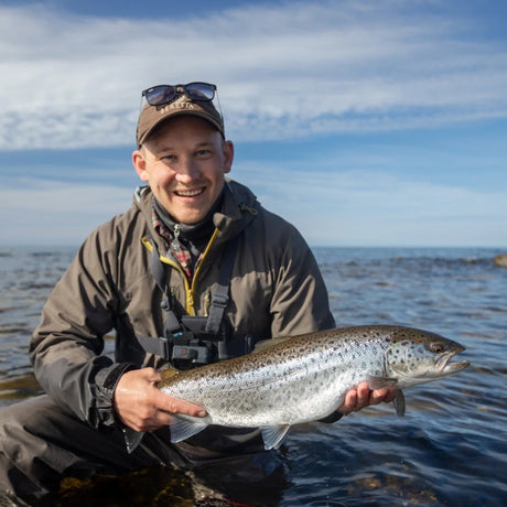 Leende sportfiskare knäböjer i vattnet och visar upp en havsöring på Gotland