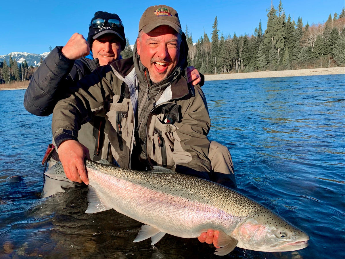 Två sportfiskare i friluftskläder knäböjer vid en älv med stor fisk, omgivna av barrträd och blå himmel under fiskeresan.