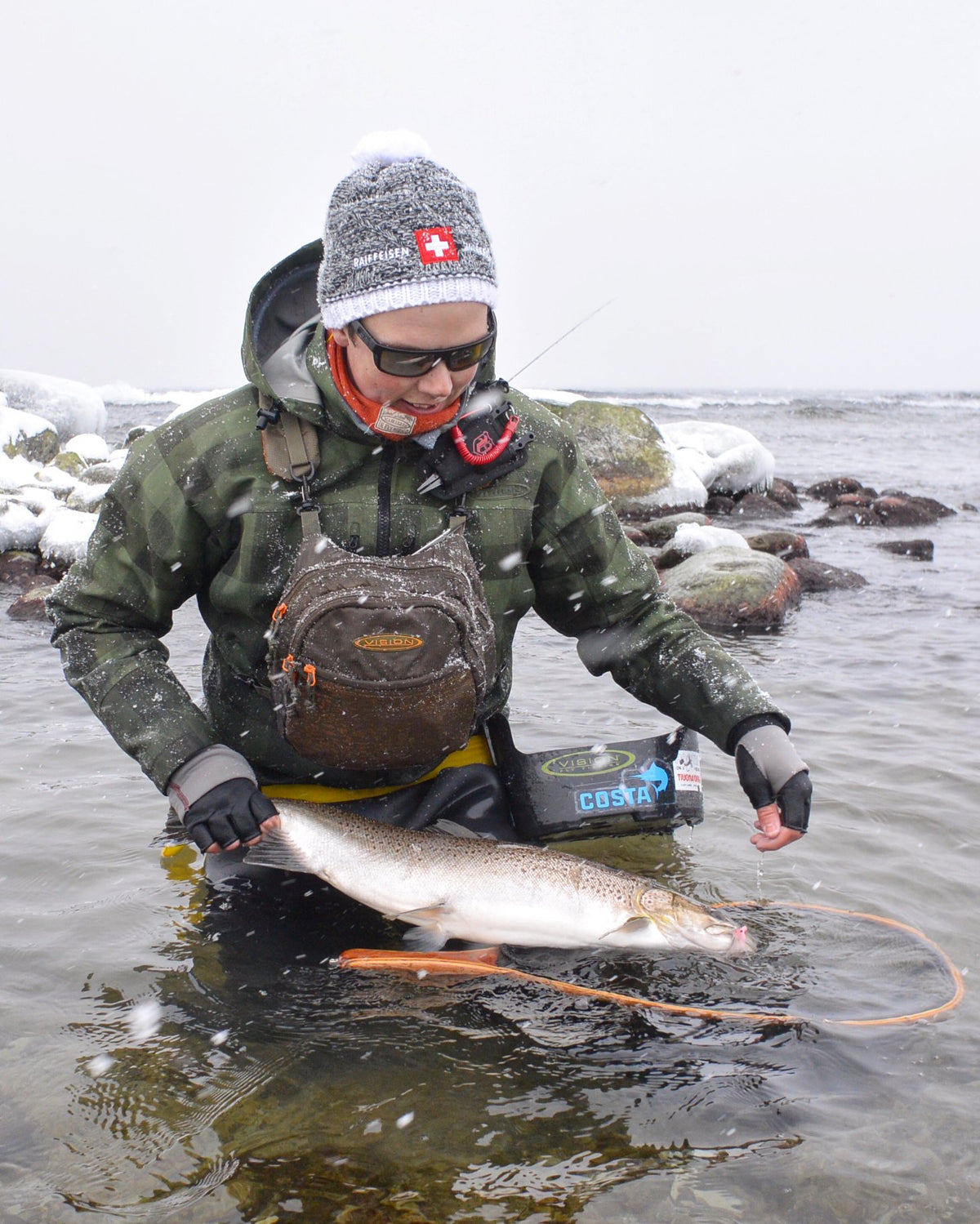 Sportfiskare i vinterkläder och grå mössa håller stor fisk i snötäckt vatten, omgiven av snöfall och stenar-drömresan fiske.