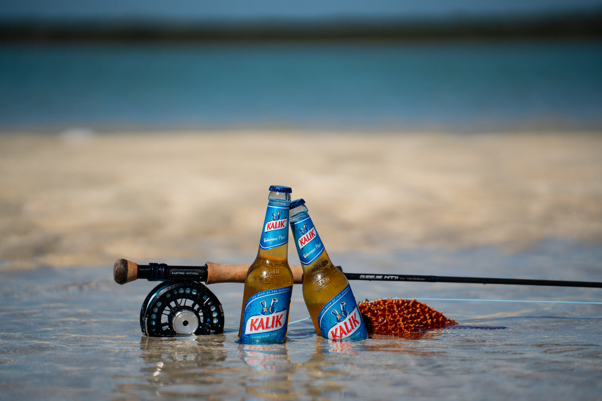 Två Kalik-ölflaskor i grunt vatten bredvid fiskespö och rulle, idealiskt för sportfiskare vid strand och blått hav.