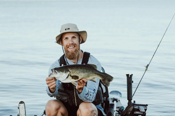 Skäggig sportfiskare med hatt sitter i båt, håller stor fisk, ler mot kameran. Lugnt vatten och fiskespö syns i bakgrunden.