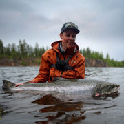 Sportfiskare med stor fångst i orange jacka vid skogskantat vatten, Fiskeresa Camp Onka Lappland.