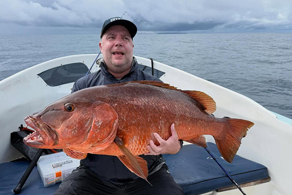 En man på en liten båt under sin fiskeresa håller en stor rödbrun fisk och ler mot kameran under molnig himmel.