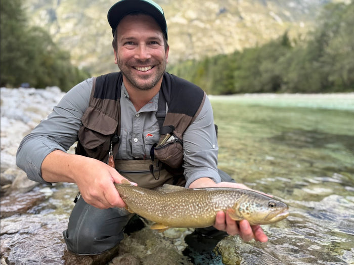 En leende sportfiskare i vadarbyxor och kepsar håller en stor öring vid en klar fjällälv, omgiven av berg och grönska.