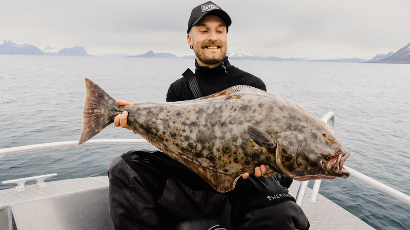 En person på en fiskebåt under en fisketur håller en stor, prickig fisk och ler. I bakgrunden syns berg och lugnt vatten under en molnig himmel.