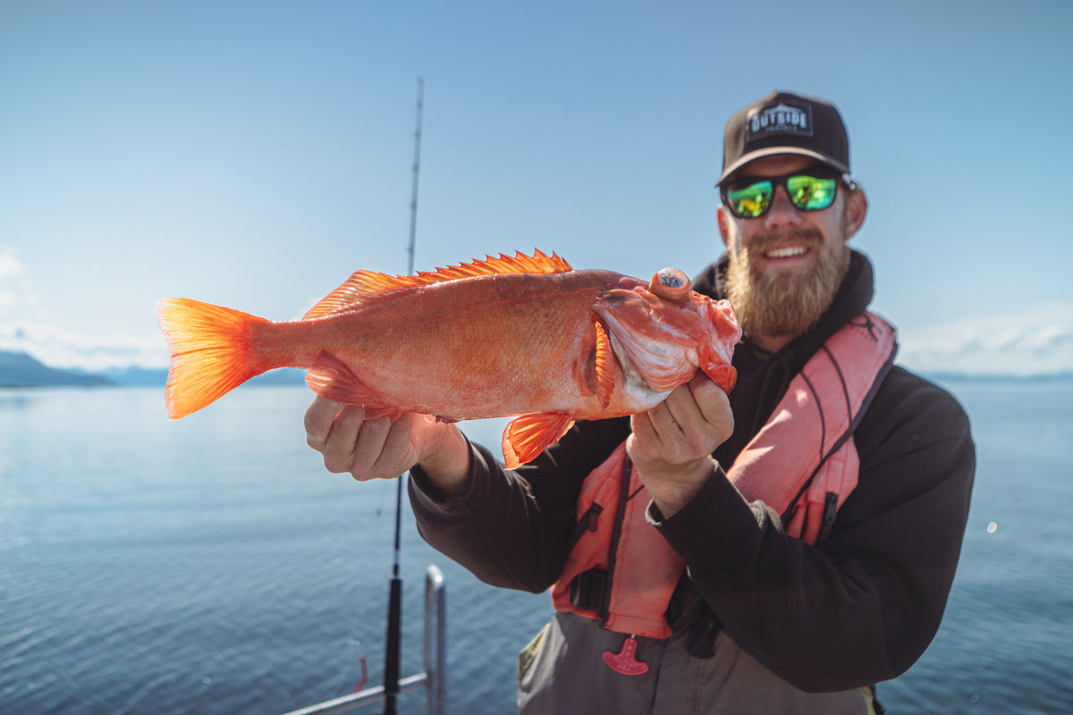 En sportfiskare med solglasögon och kepsar ler och håller upp en orange fisk på båt med lugnt vatten och klar himmel.