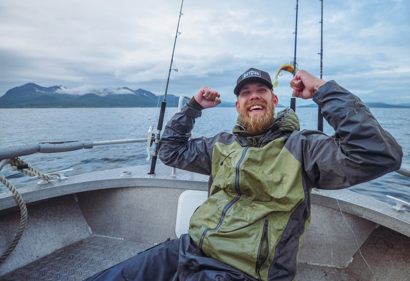 Skäggiga sportfiskare i regnjacka firar på båt med fiskespön, berg och vatten i bakgrunden - perfekt fiskeäventyr.