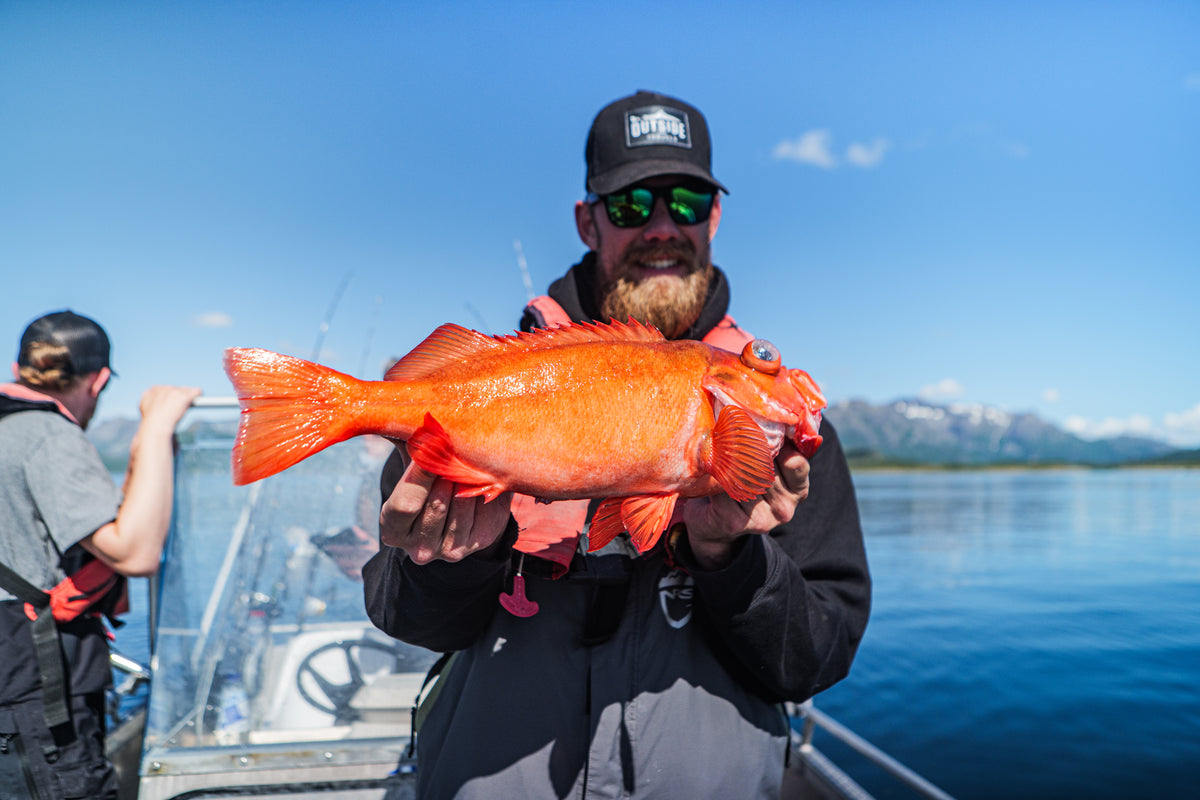 Sportfiskare med solglasögon och keps håller en stor orange fisk på båt, blått vatten och fjäll i bakgrunden under fiskeresa.
