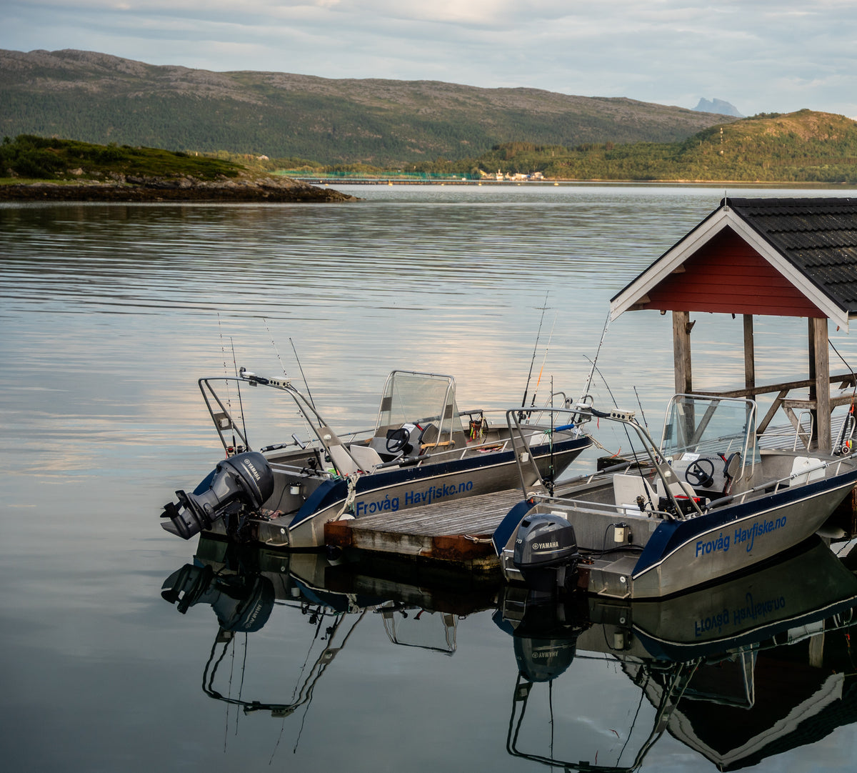 Tre små fiskebåtar med spön ligger vid ett rött trähus på lugnt vatten, gröna kullar och berg under molnig himmel.