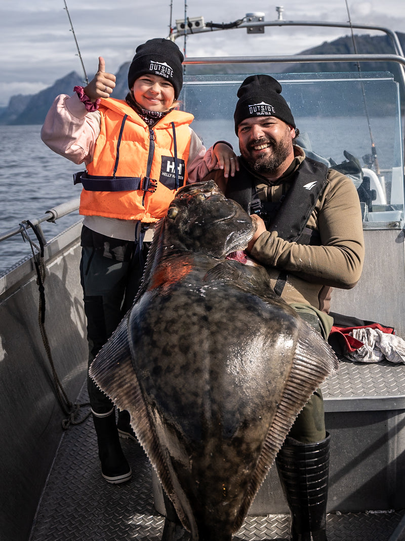 Ett leende barn och en vuxen sitter i en båt under sin fisketur, båda med svarta mössor. Barnet visar tummen upp medan den vuxne håller en stor hälleflundra i knät. I bakgrunden syns berg och vatten.
