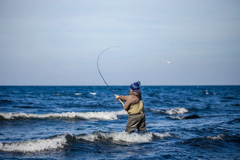 En sportfiskare i mössa, jacka och vadarbyxor fiskar i havet med spö bland små vågor under klar himmel.