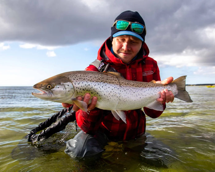 Sportfiskare i röd jacka och solglasögon knäböjer i grunt vatten med en stor, silverbrun fisk. Himlen är delvis molnig.