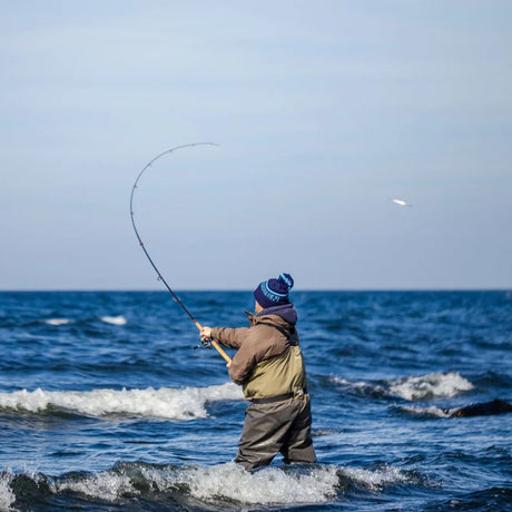 Sportfiskare i vadare och blå hatt fiskar i havet vid Gotland, kastar spinnspö under klar himmel på sportfiskeresa.