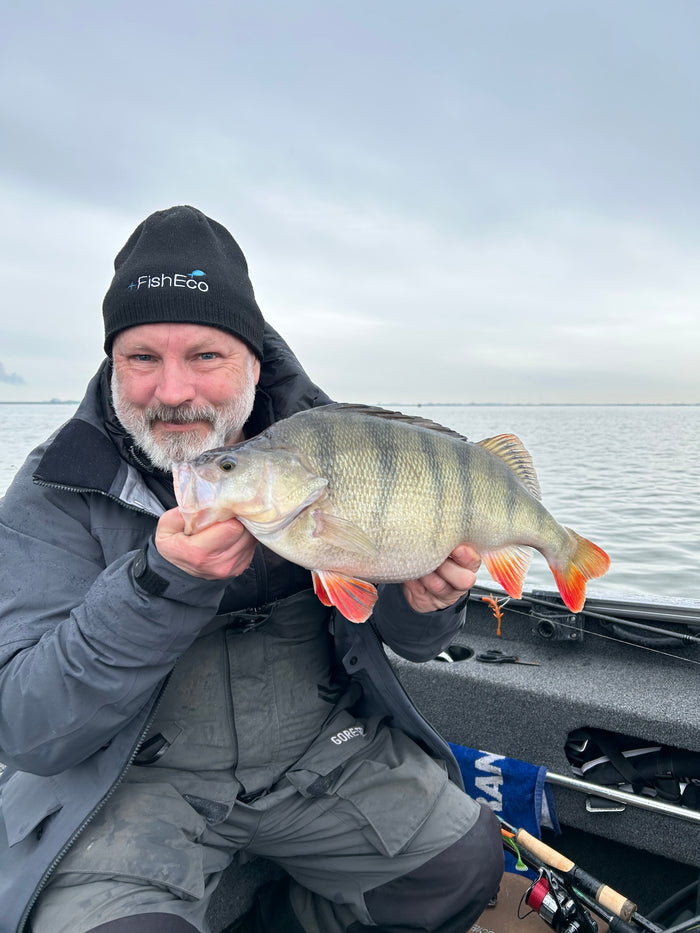 En leende sportfiskare med svart mössa visar stolt upp en stor abborre i båt på lugn sjö under fiskeresa.