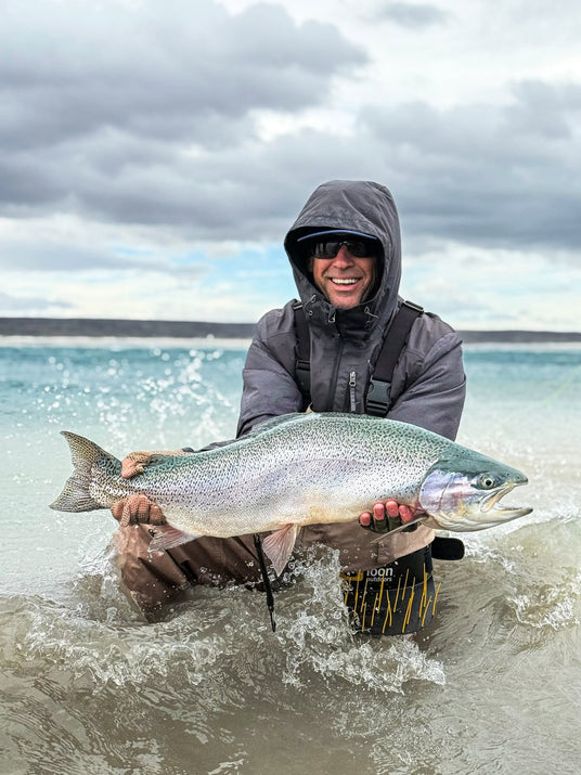 En fiskare med huva och solglasögon sitter på knä i grunt vatten och håller en stor glänsande fisk med båda händerna. Vågor slår mot stranden under en molnig himmel – ett inspirerande ögonblick från en lyckad fisketur.