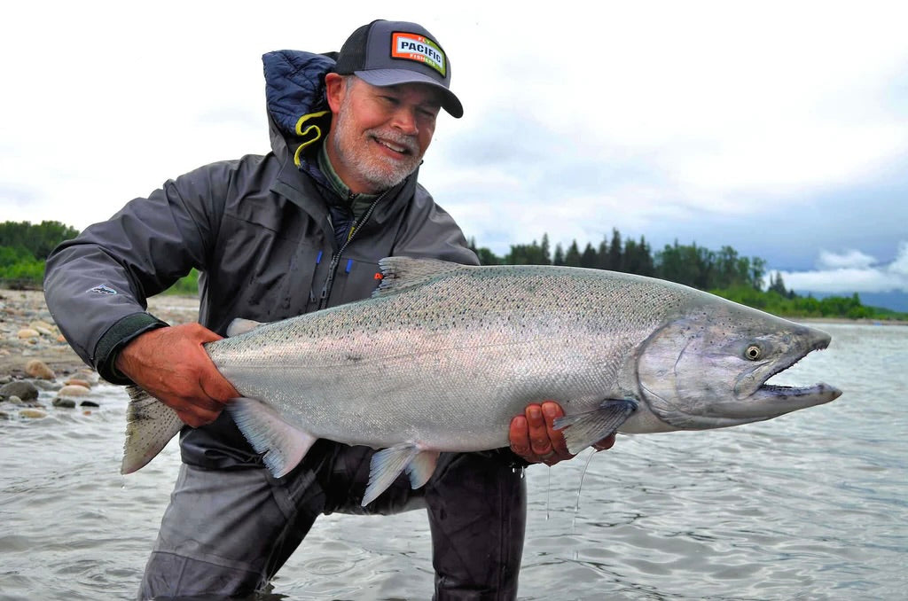 En leende man i fiskekläder sitter på knä i grunt vatten och håller upp en stor silverfisk under sin fisketur. I bakgrunden syns träd och en molnig himmel.