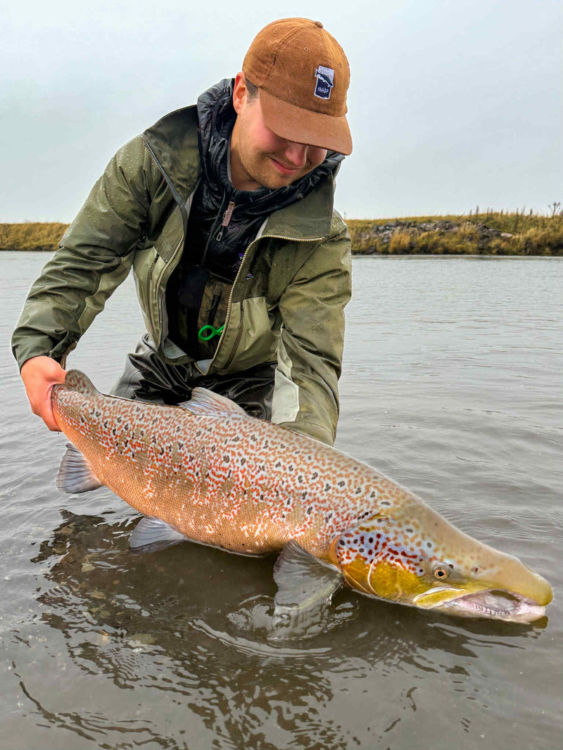 En sportfiskare i fiskeutrustning knäböjer i grunt vatten och lyfter en stor prickig fisk. Gräsbevuxen strand och molnig himmel.