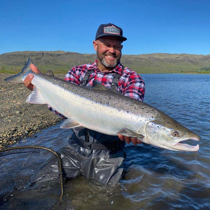 Leende sportfiskare i rutskjorta och vadare visar upp en stor lax vid glittrande vatten, redo för fiskeresa i vacker natur.