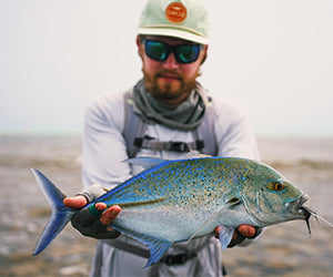 En person klädd för havsfiske - med solglasögon, hatt och långärmad skjorta - håller en blå och silverfärgad fisk med båda händerna medan han står utomhus nära vatten.