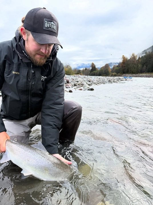 En sportfiskare i svart keps och jacka knäböjer vid en grund älv och håller varsamt en stor silverfisk nära vattenytan.