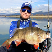 Sportfiskare med solglasögon och blå fisketröja håller stor abborrfisk vid sjö på Fiskeresa Perchzilla i Spanien.