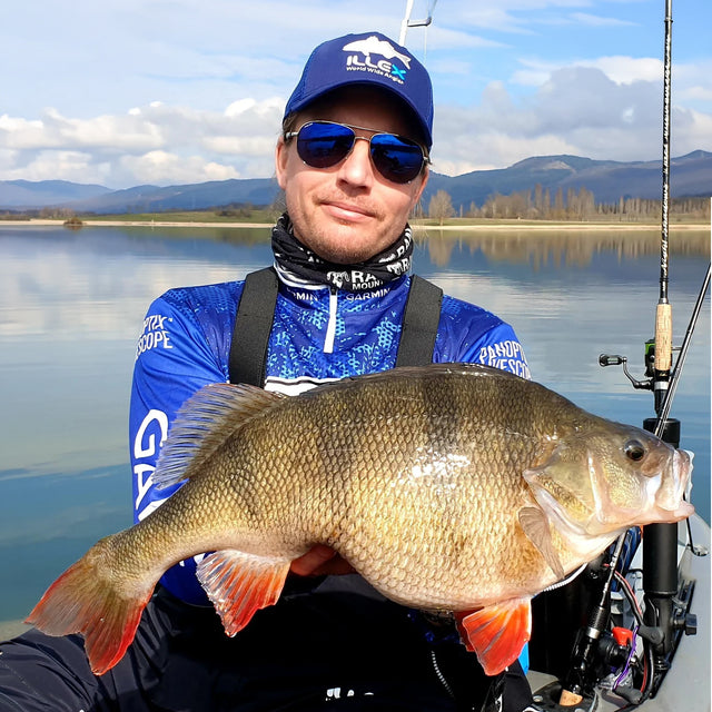Sportfiskare med solglasögon och blå fisketröja håller stor abborrfisk vid sjö på Fiskeresa Perchzilla i Spanien.