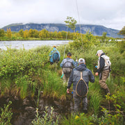Fyra sportfiskare i fiskeutrustning vandrar längs en grön älvstrand i Tjuonajokk med fjäll och skog, perfekt för fiskeresa.