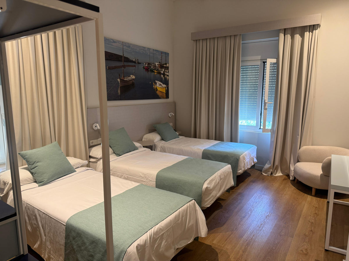 Ljust hotellrum med tre enkelsängar, sportfiskare-inspirerad inredning, trägolv, fönster, skrivbord och stol.