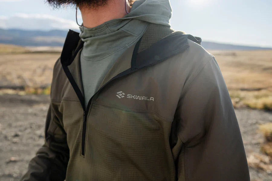 En person i grön Skwala Fishing Fusion Tactical Hoody fiskar utomhus med fält och kullar i bakgrunden.