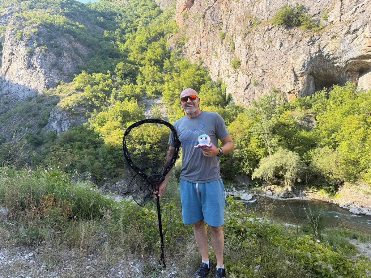 Fröhlicher Angler mit Kescher und Plüschtintenfisch vor Felsen und Fluss in Montenegro
