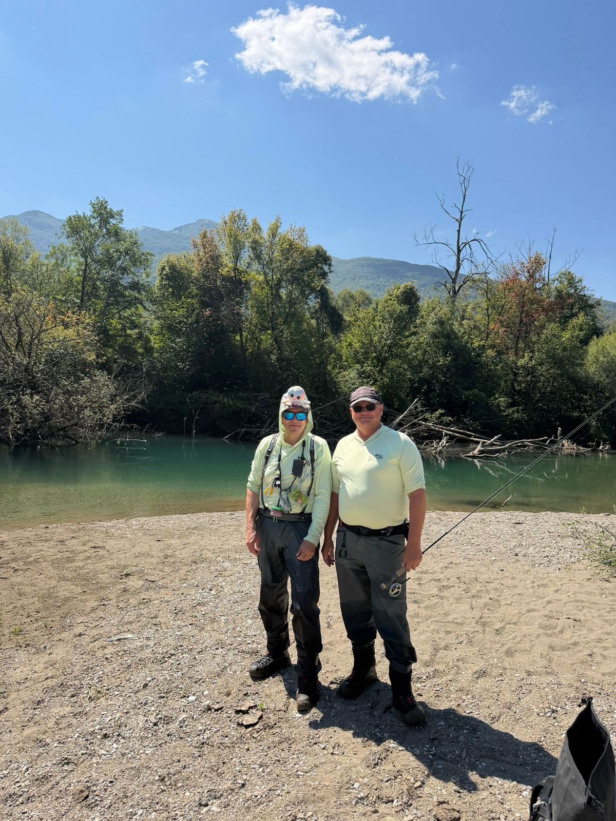 Zwei Fliegenfischer auf einer Kiesbank an einem türkisfarbenen Fluss in Montenegro, ein Plüschtintenfisch auf der Mütze des einen
