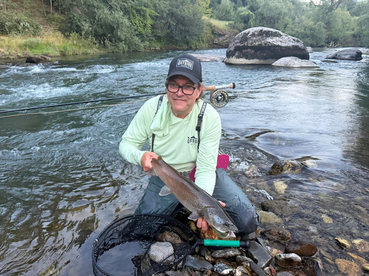Lächelnder Fliegenfischer mit großer Fisch in einem strömenden Fluss in Montenegro, mit einer Outside Travels-Kappe