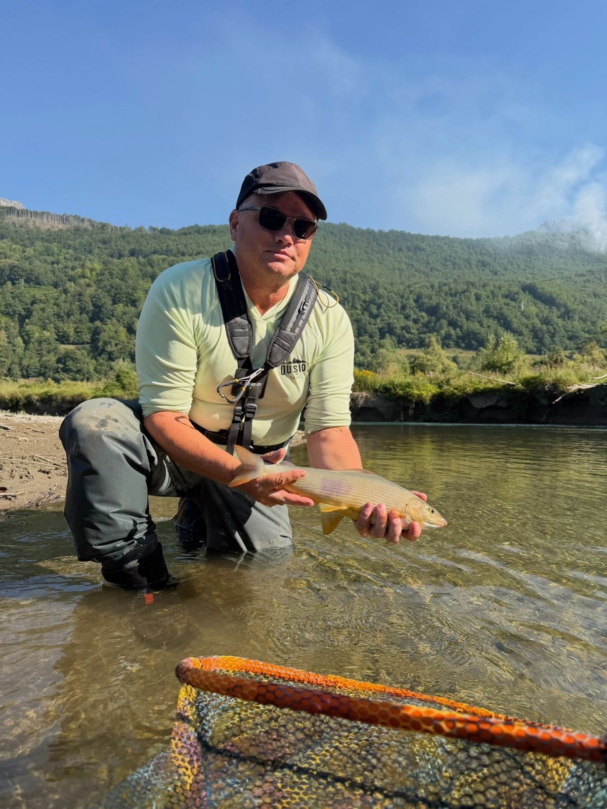 Fliegenfischer mit Äsche im Morgenlicht an einem grünen Tal in Montenegro, orangefarbener Kescher im Vordergrund